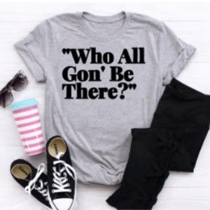 May include: A heather gray t-shirt with the text "Who All Gon' Be There?" in black. The image also shows black pants, black and white sneakers, sunglasses, and a pink and blue striped cup.