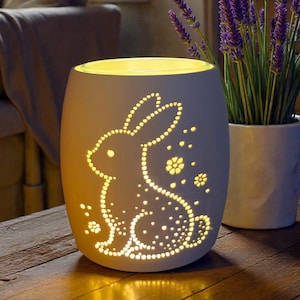 May include: A white ceramic candle holder with a cut-out bunny design, illuminated from within. The candle holder is on a wooden table, next to a potted lavender plant. The unikatHi logo is visible in the top right corner.