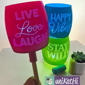 Könnte beinhalten: Eine Sammlung farbenfroher, beleuchteter Glasdekorationen. Eine wird von einer Hand gehalten und zeigt "LIVE Love LAUGH" in weißen Punkten. Andere Gläser zeigen "HAPPY Vibes" und "STAY WILD". Eine kleine Sukkulente und zwei zylindrische Objekte sind ebenfalls vorhanden.