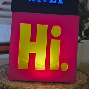 Puede incluir: Dos letreros iluminados en forma de cubo apilados. El letrero superior es negro con las palabras "GOOD VIBES ONLY" en azul. El letrero inferior es rosa con la palabra "Hi." en amarillo.