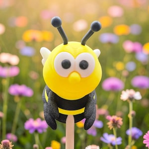 Könnte beinhalten: Ein gelb-schwarzer Gartenstecker in Bienenform mit großen Augen und Fühlern. Die Biene sitzt auf einem Holzstab, vor einem Hintergrund aus bunten Wildblumen. Das Bild enthält den Text "unikatHi".