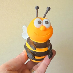 Puede incluir: Figura de abeja impresa en 3D con cabeza y cuerpo naranja, torso a rayas negras y amarillas, alas blancas y antenas negras. La abeja sonríe y tiene ojos blancos grandes. Un artículo decorativo.