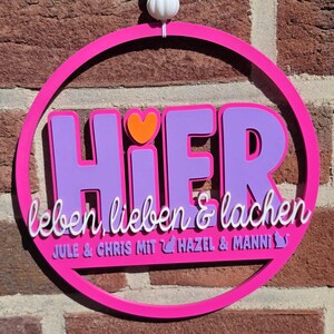 Può includere: Un cartello rotondo rosa acceso con la parola tedesca "HIER" in lettere viola, un cuore arancione e le parole "leben lieben & lachen" in corsivo bianco. Sono inclusi anche i nomi "JULE & CHRIS MIT" e "HAZEL & MANNI".