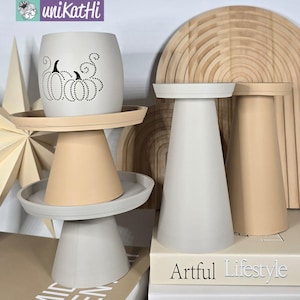 May include: A collection of decorative items, including a white pumpkin-themed planter, pedestal stands, and a wooden arched wall decor. The planter features a black pumpkin design. The stands are in neutral tones. The books say "Artful Lifestyle" and "The Art of Home."