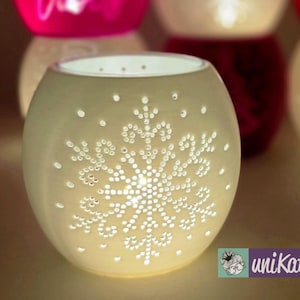 Puede incluir: Portavelas esférico color crema con diseño de copo de nieve. El copo de nieve está creado por pequeños agujeros espaciados que permiten el paso de la luz. Otros portavelas son visibles en el fondo, algunos con la palabra "Vibes".