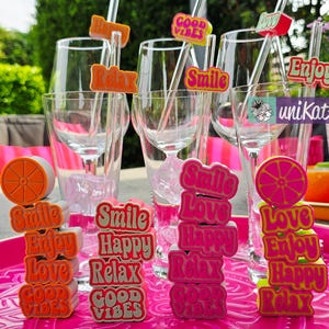 Puede incluir: Una escena al aire libre vibrante con copas de vino transparentes, pajitas y agitadores decorativos. Los agitadores presentan palabras como "Smile", "Enjoy" y "Good Vibes" en colores brillantes. Una bandeja rosa contiene pilas de las mismas palabras.