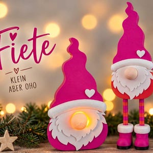 May include: Two pink wooden gnome figures with white beards and hats. One gnome is short with a glowing light, the other is tall with striped legs and boots. The text reads Fiete and KLEIN ABER OHO with heart symbols on a blurred background.