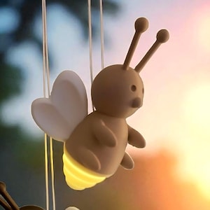 Puede incluir: Dos luces colgantes con forma de abeja, con alas blancas en forma de corazón y fondos amarillos brillantes. Una abeja es marrón, la otra es marrón claro. El fondo es una vista borrosa de una ventana con una cortina.