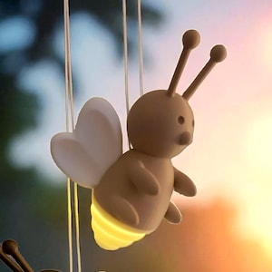 Puede incluir: Dos luces colgantes con forma de abeja, con alas blancas en forma de corazón y fondos amarillos brillantes. Una abeja es marrón, la otra es marrón claro. El fondo es una vista borrosa de una ventana con una cortina.