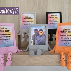 Puede incluir: Una colecci&oacute;n de marcos de fotos coloridos, cada uno con un dise&ntilde;o &uacute;nico. Los marcos son de color rosa, gris, negro y naranja, con una foto o texto que dice "Ich bin ein 'MEMORY-BUDDY'". Los marcos tienen brazos y piernas y est&aacute;n sobre una estanter&iacute;a de madera.