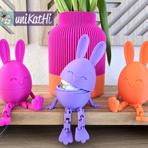 Könnte beinhalten: Drei Osterei-Behälter in Form eines Osterhasen mit Scharnierdeckeln. Die Behälter sind aus Kunststoff und in Pink, Lila und Orange lackiert. Die Behälter stehen auf einer Holzoberfläche.