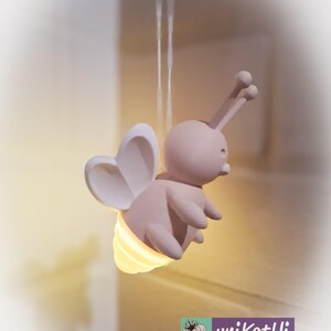 K&ouml;nnte beinhalten: Eine hellrosa, bienenf&ouml;rmige Nachtlampe mit herzf&ouml;rmigen Fl&uuml;geln und einem gelb leuchtenden Licht, das aus ihrem Hinterleib kommt. Die Lampe h&auml;ngt an einem d&uuml;nnen, wei&szlig;en Faden. Das Logo "uniKatHi" ist unten rechts zu sehen.