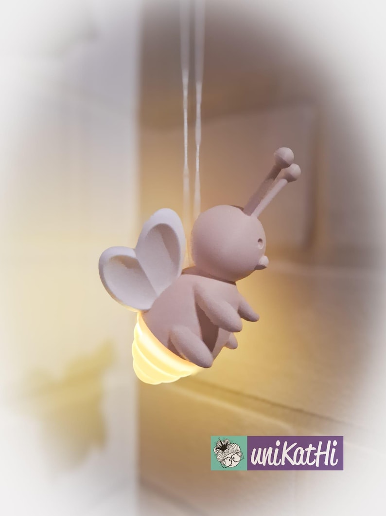 K&ouml;nnte beinhalten: Eine hellrosa, bienenf&ouml;rmige Nachtlampe mit herzf&ouml;rmigen Fl&uuml;geln und einem gelb leuchtenden Licht, das aus ihrem Hinterleib kommt. Die Lampe h&auml;ngt an einem d&uuml;nnen, wei&szlig;en Faden. Das Logo "uniKatHi" ist unten rechts zu sehen.