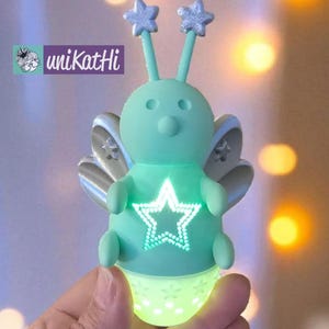 Op de afbeelding: Een mintgroen nachtlampje in de vorm van een grillige kever met stervormige antennes en vleugels. De kever heeft een oplichtende ster op zijn borst en een oplichtende basis. De merknaam "unikatHi" is zichtbaar.