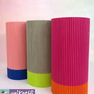 May include: Three cylindrical vases with vertical ribbed texture. The vases are in shades of pink, grey-green, and fuchsia, each with a contrasting coloured base in blue, lime green, and orange. The vases are displayed on a white surface.