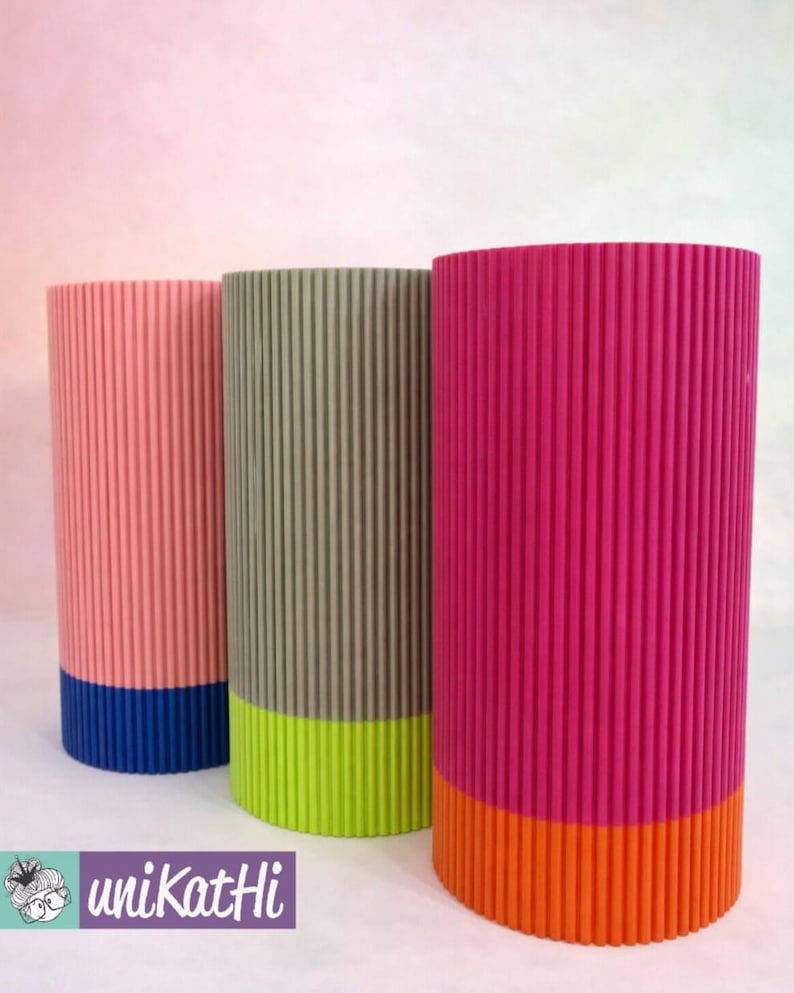 May include: Three cylindrical vases with vertical ribbed texture. The vases are in shades of pink, grey-green, and fuchsia, each with a contrasting coloured base in blue, lime green, and orange. The vases are displayed on a white surface.