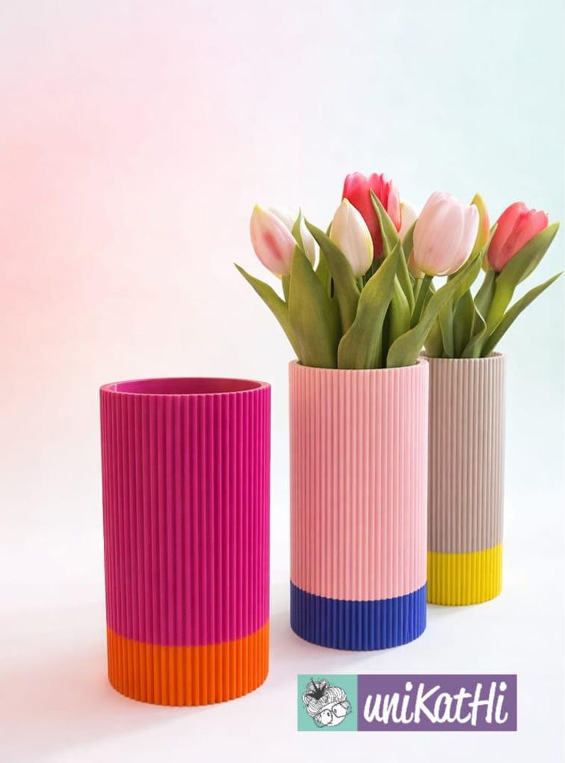 May include: Three colourful, ribbed cylindrical vases. One is magenta with an orange base, another is pink with a blue base, and the third is tan with a yellow base. The pink and tan vases hold tulips.