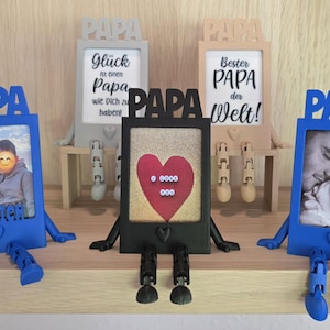 以下が含まれることがあります： 「PAPA」の文字が上部にあるフォトフレームのコレクション。フレームは青、グレー、黒など様々な色があります。写真が入っているフレームもあれば、文字が入っているフレームもあります。フレームには足と腕が付いています。
