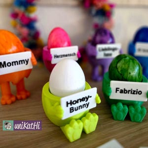 Puede incluir: Cinco soportes para huevos coloridos con nombres en ellos. Los soportes tienen forma de pequeñas personas y sostienen carteles con los nombres "Mommy", "Herzmensch", "Honey-Bunny", "Fabrizio" y "Glücklicher  Tante".