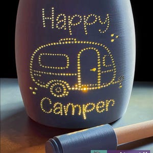 Puede incluir: Un portavelas de cerámica azul oscuro con las palabras "Happy Camper" y un contorno de caravana iluminado por pequeñas luces. Una herramienta con mango de madera descansa frente al soporte. El portavelas tiene un interior amarillo.