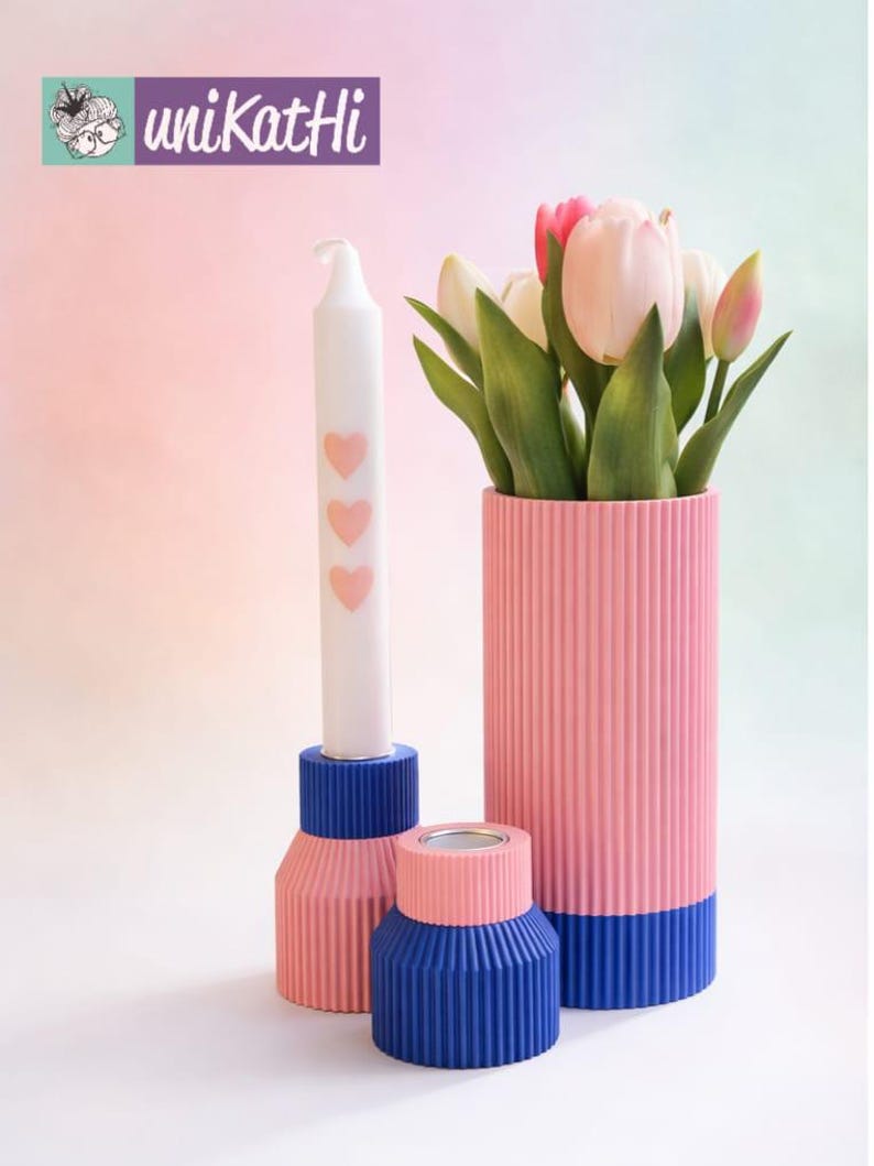 May include: A still life arrangement with a white candle adorned with pink hearts, a pink and blue vase holding tulips, and two matching candle holders. The items are in shades of pink and blue, with a ribbed texture. The logo "unikatHi" is in the upper left corner.