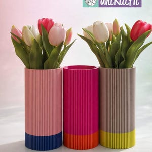 May include: Three cylindrical vases with vertical ribbed texture, each holding tulips. The vases are pink, fuchsia, and taupe, with contrasting coloured bases in blue, orange, and yellow. The top right corner has the text "uniKatHi".