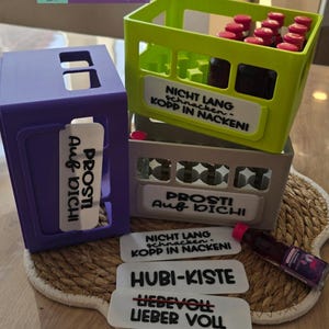 May include: A stack of three colorful miniature crates, purple, green, and gray, with white labels. The green crate holds small bottles with red caps. Text on the labels includes "PROST! Auf DICH!" and "NICHT LANG schnacken - KOPP IN NACKENI!"