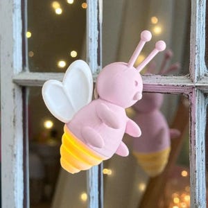 K&ouml;nnte beinhalten: Eine rosa bienenf&ouml;rmige Leuchte mit wei&szlig;en Fl&uuml;geln und gelb leuchtenden Streifen am Hinterleib. Die Biene hat zwei Antennen mit runden Spitzen. Das Licht ist an einem wei&szlig; gerahmten Fenster mit verschwommenem Hintergrund aus Lichtern befestigt.