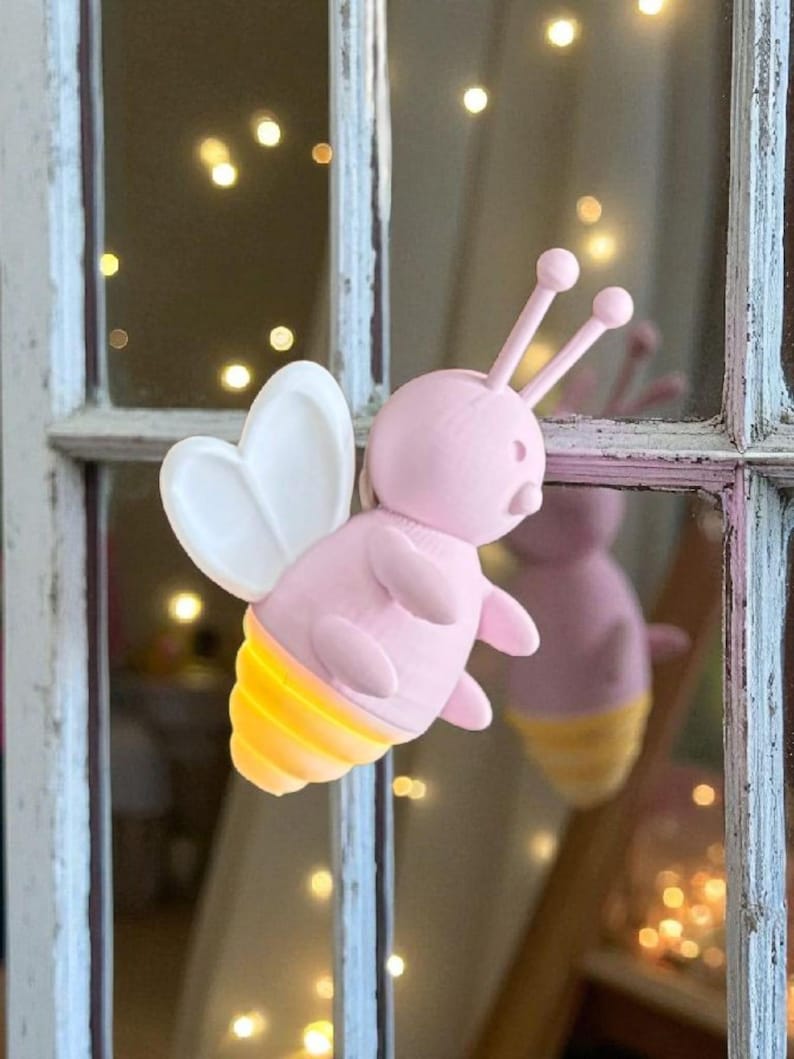 K&ouml;nnte beinhalten: Eine rosa bienenf&ouml;rmige Leuchte mit wei&szlig;en Fl&uuml;geln und gelb leuchtenden Streifen am Hinterleib. Die Biene hat zwei Antennen mit runden Spitzen. Das Licht ist an einem wei&szlig; gerahmten Fenster mit verschwommenem Hintergrund aus Lichtern befestigt.