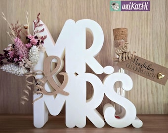 May include: White wooden "Mr. & Mrs." sign with a small glass bottle and a brown tag that says "Herzlichen Glückwunsch". The sign is decorated with dried flowers.