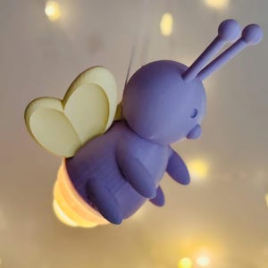 K&ouml;nnte beinhalten: Eine skurrile, lila, cartoonartige Gl&uuml;hw&uuml;rmchen-Figur mit gelben Fl&uuml;geln und einem leuchtenden, mehrfarbigen Hinterleib. Das Gl&uuml;hw&uuml;rmchen hat zwei F&uuml;hler und schwebt in der Luft. Der Hintergrund ist ein weicher, verschwommener, cremefarbener Farbton mit kleinen, unscharfen Lichtern.
