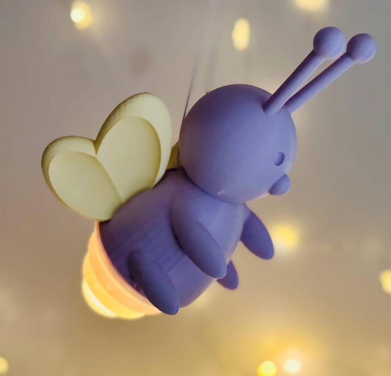 K&ouml;nnte beinhalten: Eine skurrile, lila, cartoonartige Gl&uuml;hw&uuml;rmchen-Figur mit gelben Fl&uuml;geln und einem leuchtenden, mehrfarbigen Hinterleib. Das Gl&uuml;hw&uuml;rmchen hat zwei F&uuml;hler und schwebt in der Luft. Der Hintergrund ist ein weicher, verschwommener, cremefarbener Farbton mit kleinen, unscharfen Lichtern.