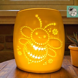 May include: A glowing yellow glass candle holder with a bee design. The bee is created with small, evenly spaced holes that allow light to shine through. A small potted succulent is next to the candle holder.