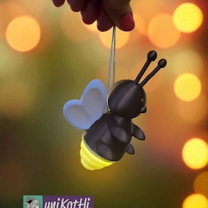 K&ouml;nnte beinhalten: Eine skurrile, dunkelbraune Biene-f&ouml;rmige Lampe mit einem leuchtend gelben Hinterleib und hellblauen Fl&uuml;geln wird an einer Schnur gehalten. Die Biene hat zwei schwarze F&uuml;hler und wird vor einem verschwommenen Hintergrund aus warmen, goldenen Bokeh-Lichtern gehalten. Das Logo "unikatHi" ist sichtbar.