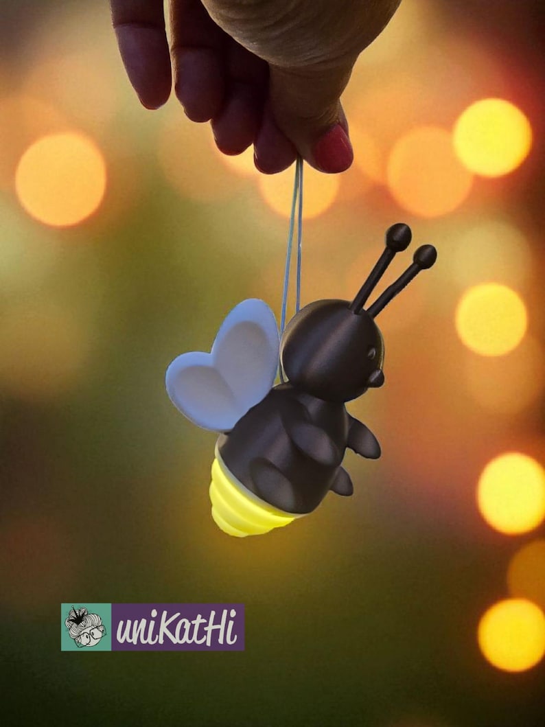 K&ouml;nnte beinhalten: Eine skurrile, dunkelbraune Biene-f&ouml;rmige Lampe mit einem leuchtend gelben Hinterleib und hellblauen Fl&uuml;geln wird an einer Schnur gehalten. Die Biene hat zwei schwarze F&uuml;hler und wird vor einem verschwommenen Hintergrund aus warmen, goldenen Bokeh-Lichtern gehalten. Das Logo "unikatHi" ist sichtbar.