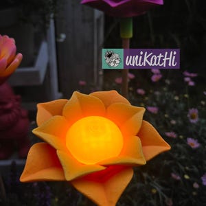 Puede incluir: Luces solares con forma de flor naranja y morada con centros brillantes. Los p&eacute;talos de las flores son detallados y las luces est&aacute;n sobre estacas de madera. Incluye el texto "uniKatHi".