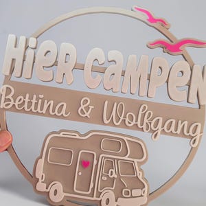 Puede incluir: Un letrero de madera con las palabras "Hier campen" y los nombres "Bettina & Wolfgang". El letrero presenta una autocaravana con un corazón rosa y dos pájaros rosas. El letrero tiene forma circular.
