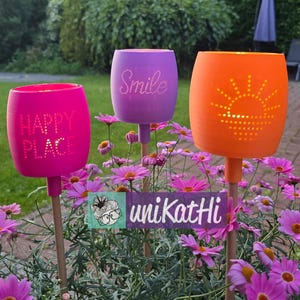 Può includere: Tre luci da giardino colorate, rosa, viola e arancione. La luce rosa dice "HAPPY PLACE", quella viola "Smile", e quella arancione ha un disegno di sole. Sono poste tra fiori rosa in un giardino.