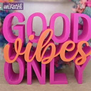 Op de afbeelding: Een roze en oranje houten bord met de woorden "Good Vibes Only" in een vet, decoratief lettertype.
