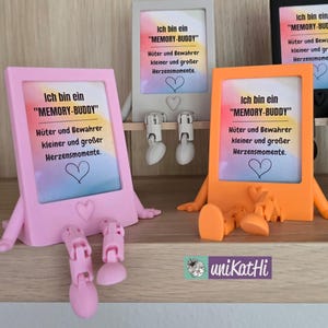 May include: A display of colorful, whimsical picture frames, each with a unique design. The frames are in pink, gray, and orange, with attached legs and arms. Each frame has the text "MEMORY-BUDDY" and a heart symbol. The frames are on a wooden shelf.