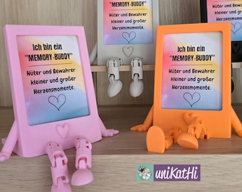 May include: A display of colorful, whimsical picture frames, each with a unique design. The frames are in pink, gray, and orange, with attached legs and arms. Each frame has the text "MEMORY-BUDDY" and a heart symbol. The frames are on a wooden shelf.