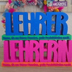Op de afbeelding: Een blauw en roze decoratief bord met de woorden "LEHRER" en "LEHRERIN" op elkaar gestapeld. Het bord bevat ook de tekst "Person, die aus kleinen Menschen, große Persönlichkeiten macht."