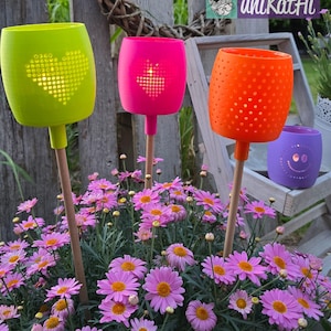 Könnte beinhalten: Helle Gartenlaternen in Grün, Pink und Orange, jede mit einem einzigartigen Lochmuster. Die Laternen sind auf Holzstäben montiert und zwischen rosa Gänseblümchen platziert. Eine lila Laterne mit einem Smiley ist ebenfalls zu sehen. Der Text "unikatHi" ist enthalten.
