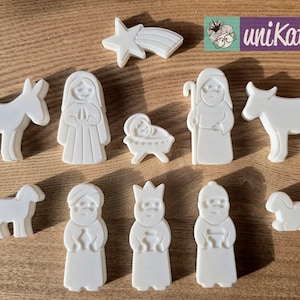 Könnte beinhalten: Eine Sammlung weißer Keramik-Weihnachtskrippenfiguren, darunter ein Stern, ein Esel, Maria, Jesuskind in einer Krippe, Josef, zwei Lämmer und drei Weise. Die Figuren sind auf einer Holzoberfläche angeordnet.