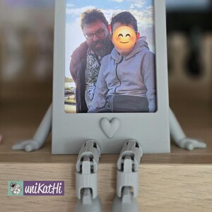 Puede incluir: Un marco de fotos gris con una foto de dos personas, con brazos y piernas adjuntos. El marco tiene un detalle de coraz&oacute;n y est&aacute; colocado sobre una superficie de madera. Las patas del marco est&aacute;n dobladas, como si estuviera sentado.