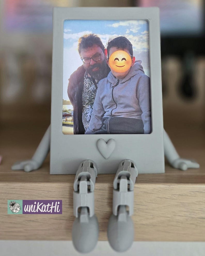 Puede incluir: Un marco de fotos gris con una foto de dos personas, con brazos y piernas adjuntos. El marco tiene un detalle de coraz&oacute;n y est&aacute; colocado sobre una superficie de madera. Las patas del marco est&aacute;n dobladas, como si estuviera sentado.