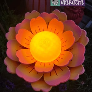 Puede incluir: Una gran escultura de flores iluminada con p&eacute;talos en capas en tonos naranja y rosa. El centro brilla con una c&aacute;lida luz amarilla. La escultura est&aacute; al aire libre, con un fondo borroso.