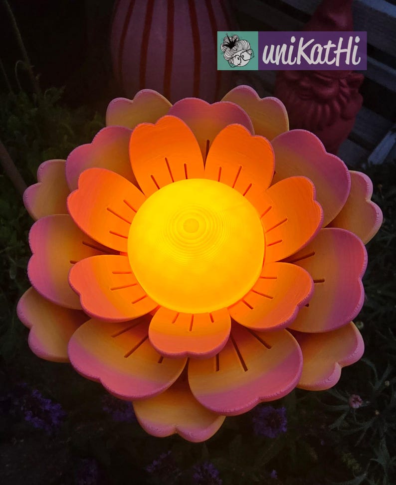 Puede incluir: Una gran escultura de flores iluminada con p&eacute;talos en capas en tonos naranja y rosa. El centro brilla con una c&aacute;lida luz amarilla. La escultura est&aacute; al aire libre, con un fondo borroso.