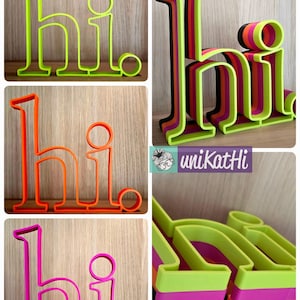 May include: Four different colored 3D printed "hi" signs. The signs are layered and made of green, orange, pink, and black plastic.