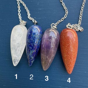 May include: Four gemstone pendulums with silver chains. The pendulums are numbered 1 through 4. The gemstones are clear, blue, purple, and orange. The pendulums are teardrop-shaped and hang against a blue background.